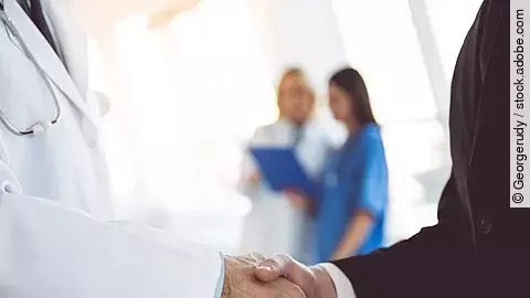 Ein Arzt und ein Geschäftsmann schütteln Hände. Im Hintergrund sind zwei Krankenhausmitarbeiterinnen verschwommen zu sehen, eine Ärztin und eine Pflegerin.