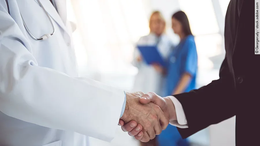 Ein Arzt und ein Geschäftsmann schütteln Hände. Im Hintergrund sind zwei Krankenhausmitarbeiterinnen verschwommen zu sehen, eine Ärztin und eine Pflegerin.
