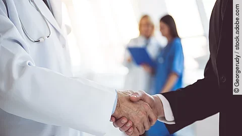 Ein Arzt und ein Geschäftsmann schütteln Hände. Im Hintergrund sind zwei Krankenhausmitarbeiterinnen verschwommen zu sehen, eine Ärztin und eine Pflegerin.