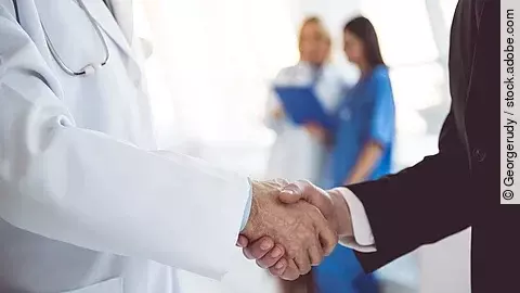 Ein Arzt und ein Geschäftsmann schütteln Hände. Im Hintergrund sind zwei Krankenhausmitarbeiterinnen verschwommen zu sehen, eine Ärztin und eine Pflegerin.