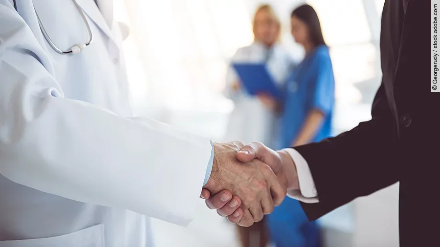 Händeschütteln Ein Arzt und ein Geschäftsmann schütteln Hände. Im Hintergrund sind zwei Krankenhausmitarbeiterinnen verschwommen zu sehen, eine Ärztin und eine Pflegerin.