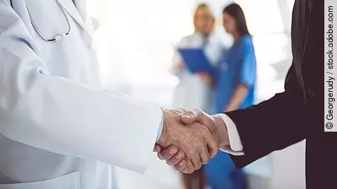 Ein Arzt und ein Geschäftsmann schütteln Hände. Im Hintergrund sind zwei Krankenhausmitarbeiterinnen verschwommen zu sehen, eine Ärztin und eine Pflegerin.