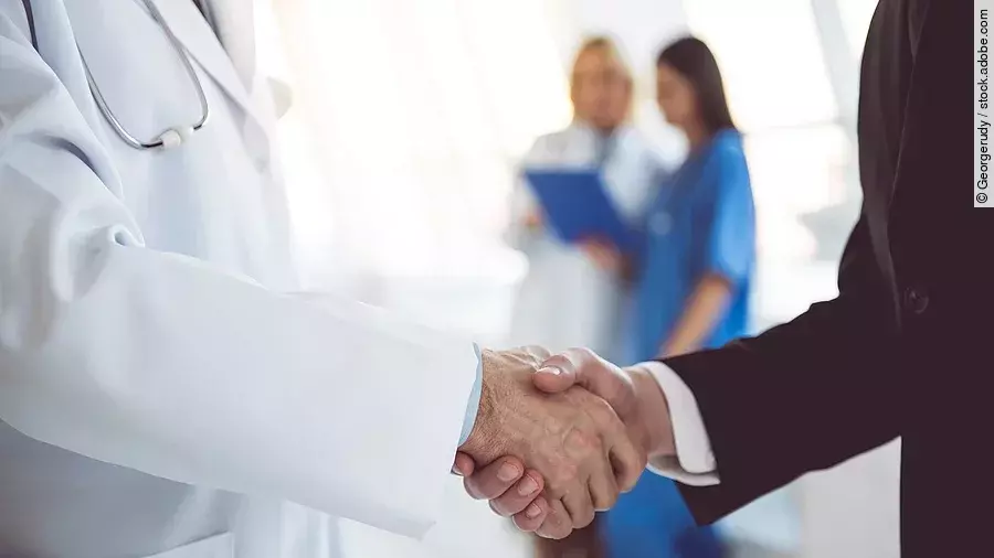 Ein Arzt und ein Geschäftsmann schütteln Hände. Im Hintergrund sind zwei Krankenhausmitarbeiterinnen verschwommen zu sehen, eine Ärztin und eine Pflegerin.