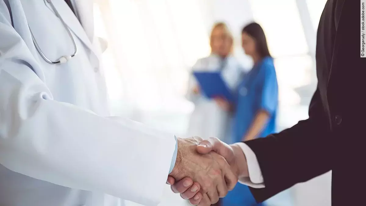 Ein Arzt und ein Geschäftsmann schütteln Hände. Im Hintergrund sind zwei Krankenhausmitarbeiterinnen verschwommen zu sehen, eine Ärztin und eine Pflegerin.
