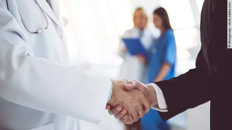 Ein Arzt und ein Geschäftsmann schütteln Hände. Im Hintergrund sind zwei Krankenhausmitarbeiterinnen verschwommen zu sehen, eine Ärztin und eine Pflegerin.