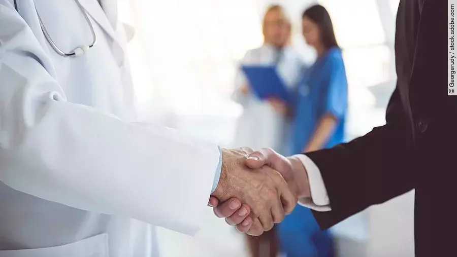 Händeschütteln Ein Arzt und ein Geschäftsmann schütteln Hände. Im Hintergrund sind zwei Krankenhausmitarbeiterinnen verschwommen zu sehen, eine Ärztin und eine Pflegerin.