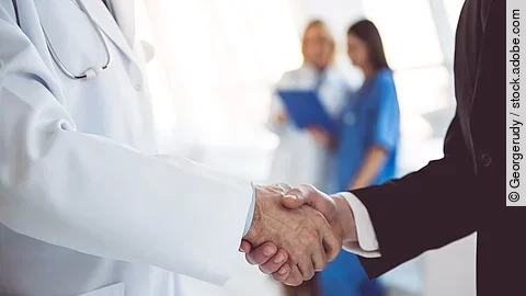 Händeschütteln Ein Arzt und ein Geschäftsmann schütteln Hände. Im Hintergrund sind zwei Krankenhausmitarbeiterinnen verschwommen zu sehen, eine Ärztin und eine Pflegerin.