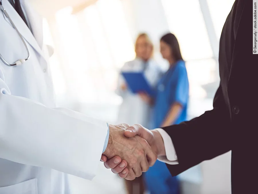 Ein Arzt und ein Geschäftsmann schütteln Hände. Im Hintergrund sind zwei Krankenhausmitarbeiterinnen verschwommen zu sehen, eine Ärztin und eine Pflegerin.