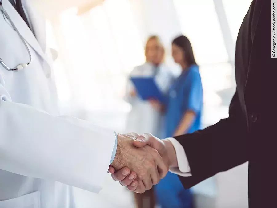 Ein Arzt und ein Geschäftsmann schütteln Hände. Im Hintergrund sind zwei Krankenhausmitarbeiterinnen verschwommen zu sehen, eine Ärztin und eine Pflegerin.