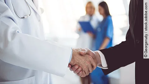 Ein Arzt und ein Geschäftsmann schütteln Hände. Im Hintergrund sind zwei Krankenhausmitarbeiterinnen verschwommen zu sehen, eine Ärztin und eine Pflegerin.