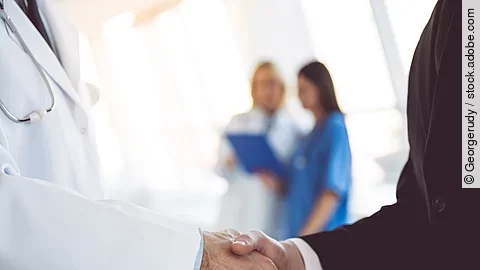 Ein Arzt und ein Geschäftsmann schütteln Hände. Im Hintergrund sind zwei Krankenhausmitarbeiterinnen verschwommen zu sehen, eine Ärztin und eine Pflegerin.