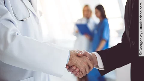 Ein Arzt und ein Geschäftsmann schütteln Hände. Im Hintergrund sind zwei Krankenhausmitarbeiterinnen verschwommen zu sehen, eine Ärztin und eine Pflegerin.