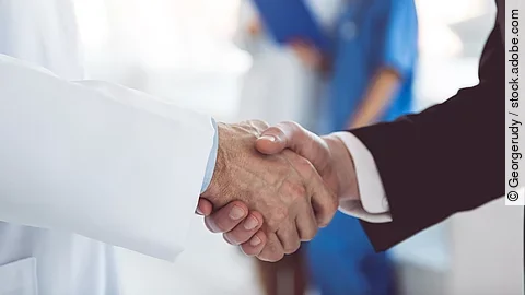 Ein Arzt und ein Geschäftsmann schütteln Hände. Im Hintergrund sind zwei Krankenhausmitarbeiterinnen verschwommen zu sehen, eine Ärztin und eine Pflegerin.