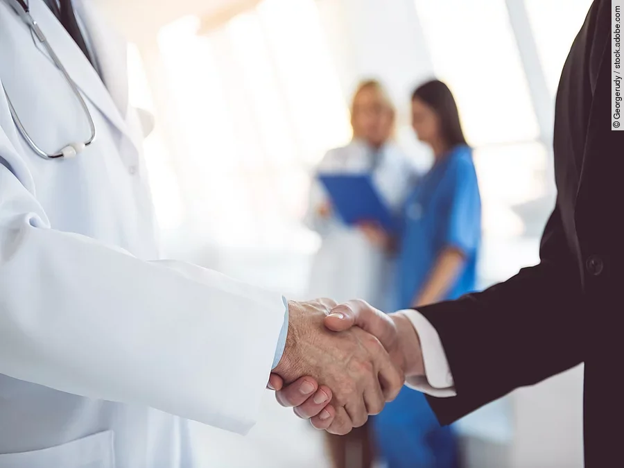 Händeschütteln Ein Arzt und ein Geschäftsmann schütteln Hände. Im Hintergrund sind zwei Krankenhausmitarbeiterinnen verschwommen zu sehen, eine Ärztin und eine Pflegerin.