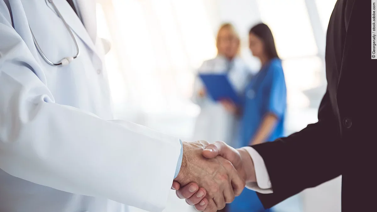 Ein Arzt und ein Geschäftsmann schütteln Hände. Im Hintergrund sind zwei Krankenhausmitarbeiterinnen verschwommen zu sehen, eine Ärztin und eine Pflegerin.