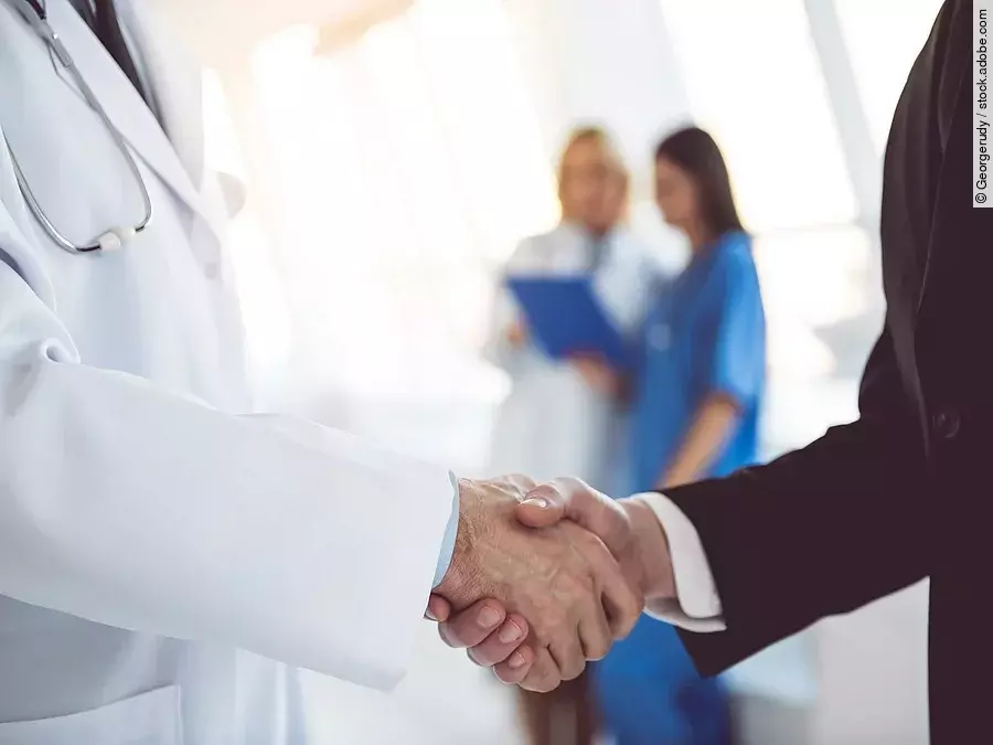 Ein Arzt und ein Geschäftsmann schütteln Hände. Im Hintergrund sind zwei Krankenhausmitarbeiterinnen verschwommen zu sehen, eine Ärztin und eine Pflegerin.