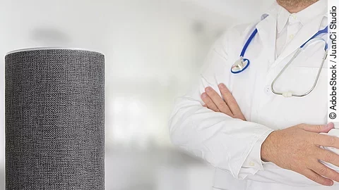 Sprachassistent Alexa Sprachassistent Alexa steht auf weißem Untergrund. Dahinter wendet sich ein Arzt mit verschränkten Armen dieser Box zu.