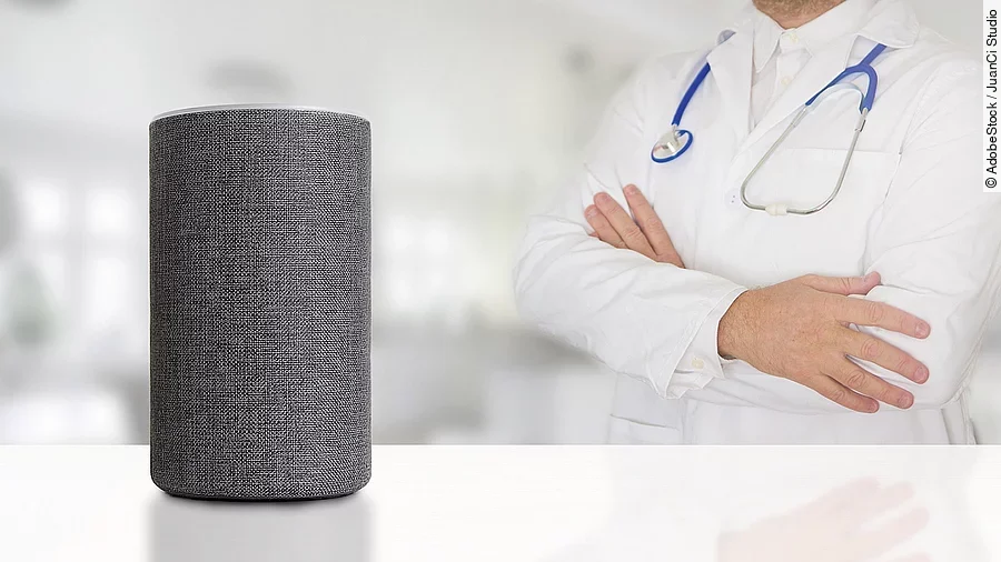 Sprachassistent Alexa steht auf weißem Untergrund. Dahinter wendet sich ein Arzt mit verschränkten Armen dieser Box zu.