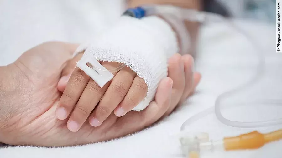 Eine Hand mit einem Verband und einer Infusion auf einem Krankenbett. Sie wird von einer anderen Hand gehalten.