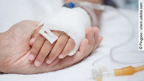 Krankenpflege Eine Hand mit einem Verband und einer Infusion auf einem Krankenbett. Sie wird von einer anderen Hand gehalten.