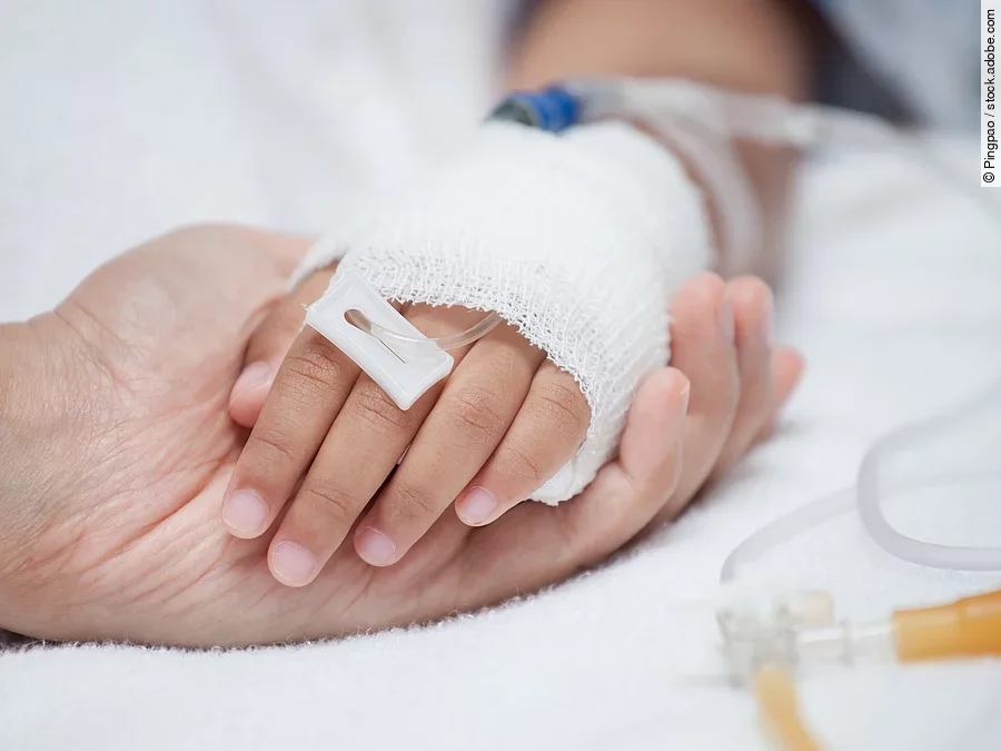 Krankenpflege Eine Hand mit einem Verband und einer Infusion auf einem Krankenbett. Sie wird von einer anderen Hand gehalten.