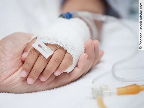 Eine Hand mit einem Verband und einer Infusion auf einem Krankenbett. Sie wird von einer anderen Hand gehalten.