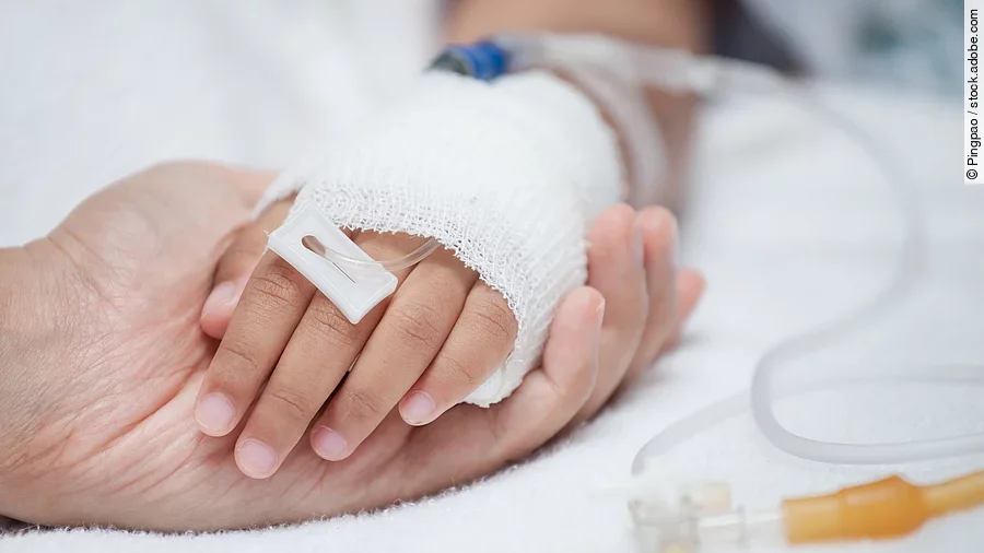 Eine Hand mit einem Verband und einer Infusion auf einem Krankenbett. Sie wird von einer anderen Hand gehalten.