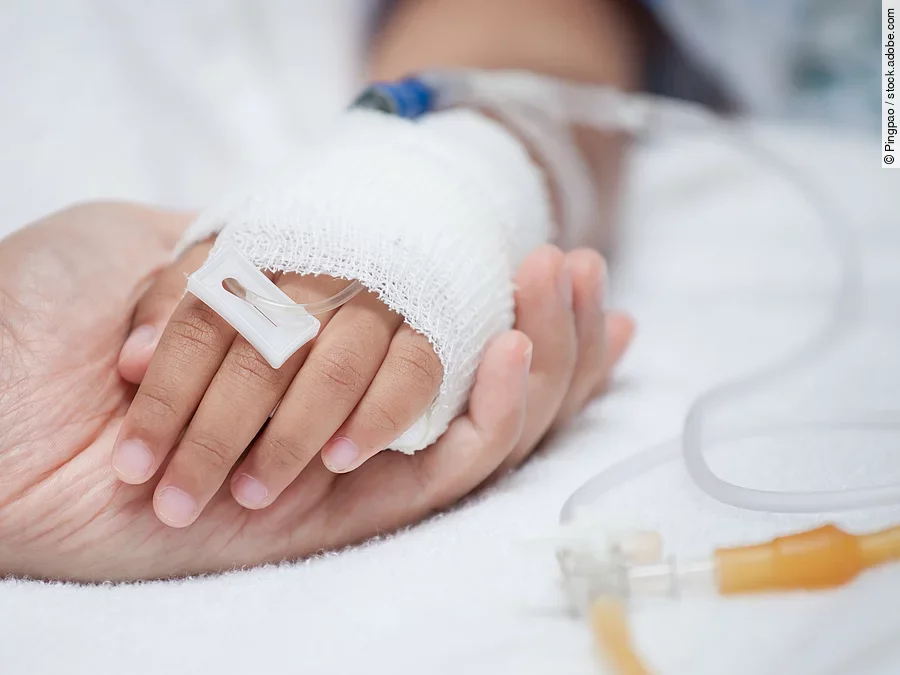 Eine Hand mit einem Verband und einer Infusion auf einem Krankenbett. Sie wird von einer anderen Hand gehalten.