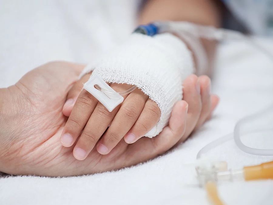 Krankenpflege Eine Hand mit einem Verband und einer Infusion auf einem Krankenbett. Sie wird von einer anderen Hand gehalten.