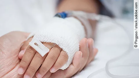 Eine Hand mit einem Verband und einer Infusion auf einem Krankenbett. Sie wird von einer anderen Hand gehalten.