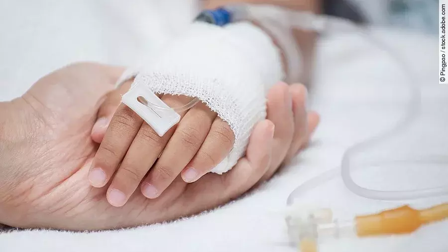 Eine Hand mit einem Verband und einer Infusion auf einem Krankenbett. Sie wird von einer anderen Hand gehalten.