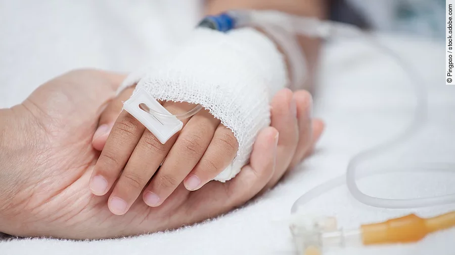 Eine Hand mit einem Verband und einer Infusion auf einem Krankenbett. Sie wird von einer anderen Hand gehalten.