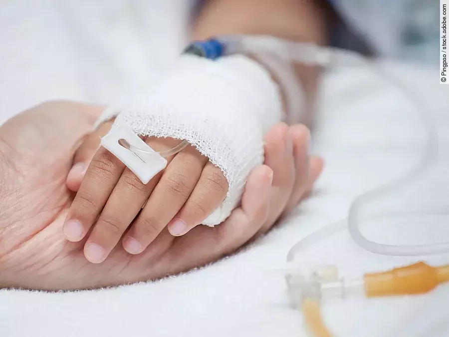 Eine Hand mit einem Verband und einer Infusion auf einem Krankenbett. Sie wird von einer anderen Hand gehalten.