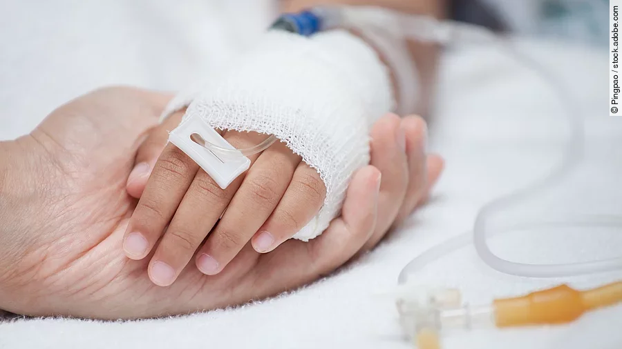 Eine Hand mit einem Verband und einer Infusion auf einem Krankenbett. Sie wird von einer anderen Hand gehalten.