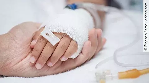 Eine Hand mit einem Verband und einer Infusion auf einem Krankenbett. Sie wird von einer anderen Hand gehalten.