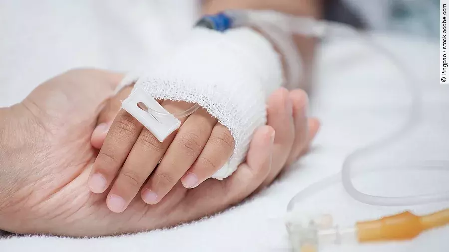 Krankenpflege Eine Hand mit einem Verband und einer Infusion auf einem Krankenbett. Sie wird von einer anderen Hand gehalten.