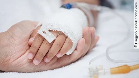 Krankenpflege Eine Hand mit einem Verband und einer Infusion auf einem Krankenbett. Sie wird von einer anderen Hand gehalten.