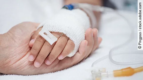 Eine Hand mit einem Verband und einer Infusion auf einem Krankenbett. Sie wird von einer anderen Hand gehalten.