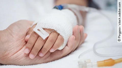 Eine Hand mit einem Verband und einer Infusion auf einem Krankenbett. Sie wird von einer anderen Hand gehalten.