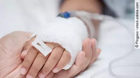 Krankenpflege Eine Hand mit einem Verband und einer Infusion auf einem Krankenbett. Sie wird von einer anderen Hand gehalten.