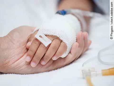 Eine Hand mit einem Verband und einer Infusion auf einem Krankenbett. Sie wird von einer anderen Hand gehalten.