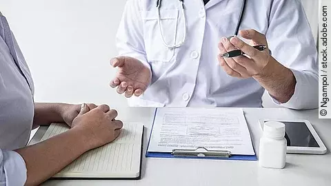 Arzt mit Patient Arzt sitzt seiner Patienten gegenüber und erklärt. Auf dem Tisch vor ihm liegen Dokumente und ein Wasserglas.