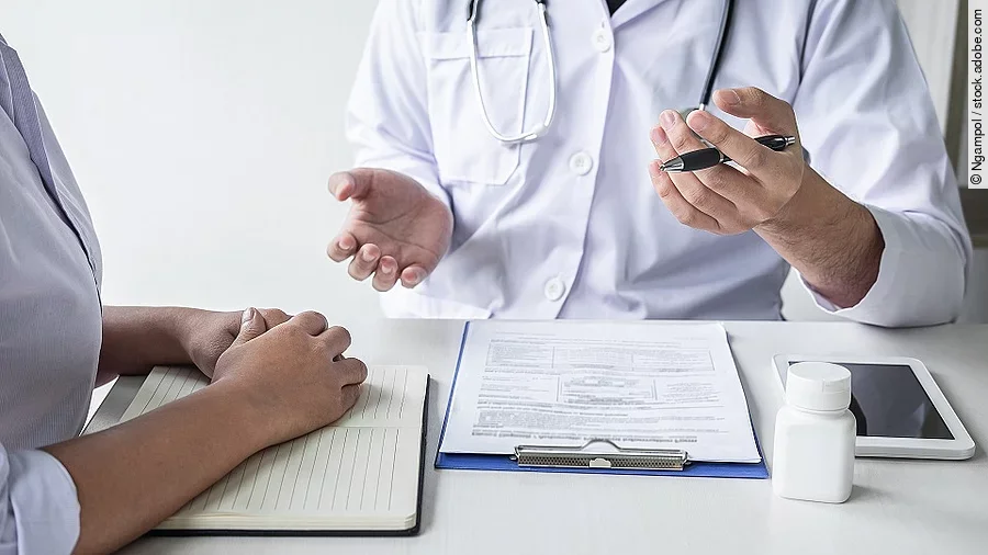 Arzt sitzt seiner Patienten gegenüber und erklärt. Auf dem Tisch vor ihm liegen Dokumente und ein Wasserglas.