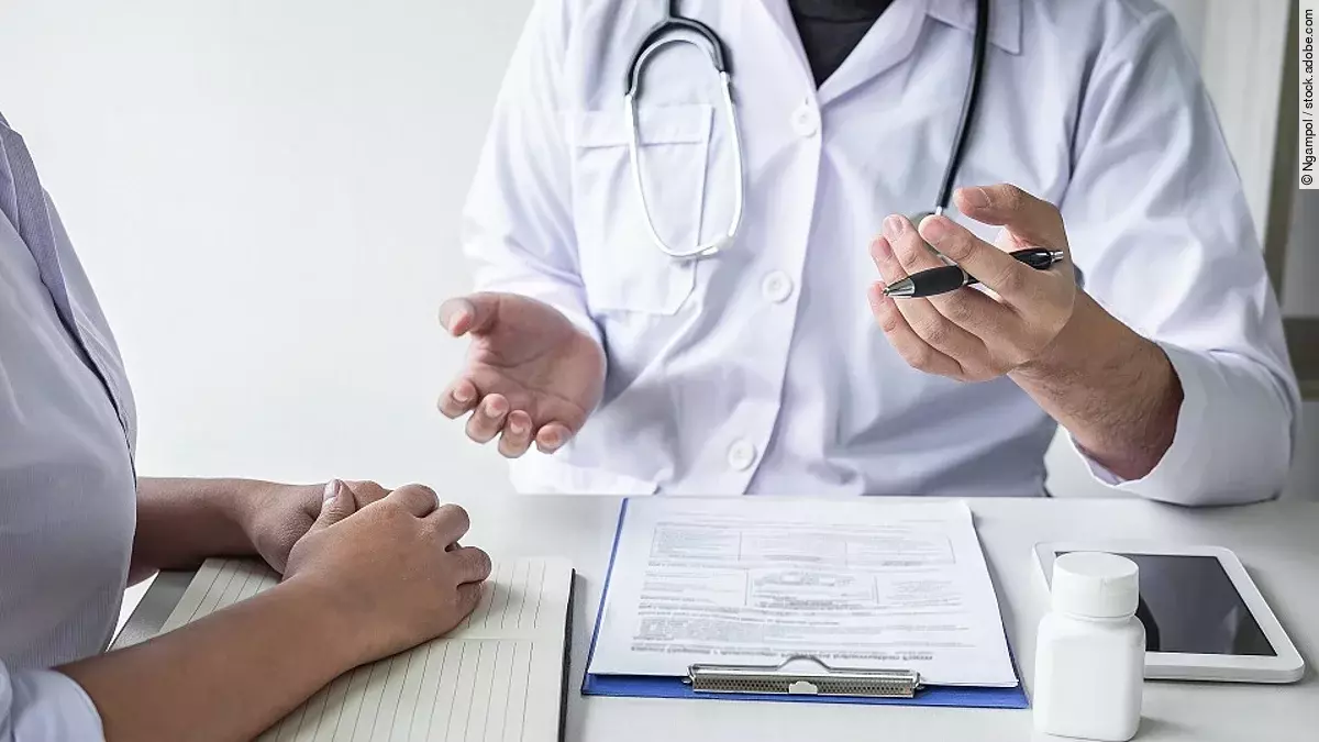 Arzt sitzt seiner Patienten gegenüber und erklärt. Auf dem Tisch vor ihm liegen Dokumente und ein Wasserglas.