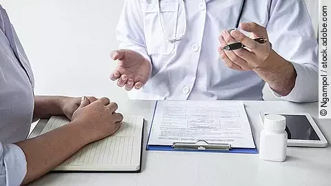 Arzt sitzt seiner Patienten gegenüber und erklärt. Auf dem Tisch vor ihm liegen Dokumente und ein Wasserglas.