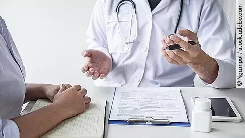 Arzt sitzt seiner Patienten gegenüber und erklärt. Auf dem Tisch vor ihm liegen Dokumente und ein Wasserglas.