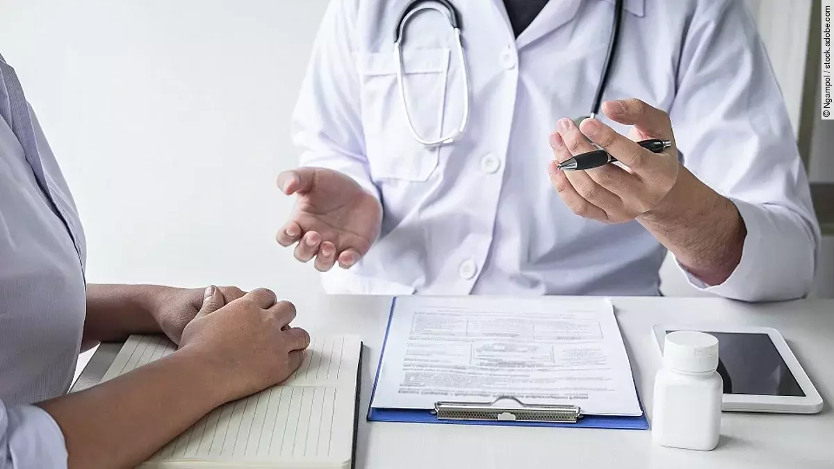 Arzt sitzt seiner Patienten gegenüber und erklärt. Auf dem Tisch vor ihm liegen Dokumente und ein Wasserglas.