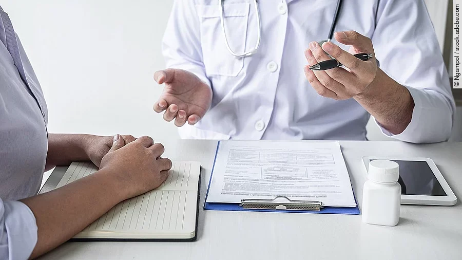 Arzt mit Patient Arzt sitzt seiner Patienten gegenüber und erklärt. Auf dem Tisch vor ihm liegen Dokumente und ein Wasserglas.