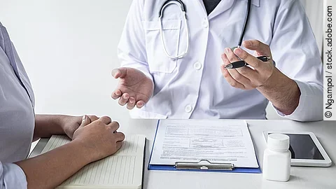 Arzt sitzt seiner Patienten gegenüber und erklärt. Auf dem Tisch vor ihm liegen Dokumente und ein Wasserglas.