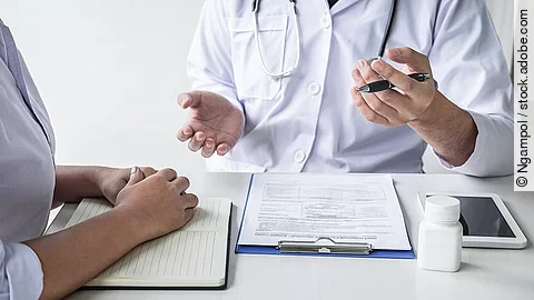 Arzt sitzt seiner Patienten gegenüber und erklärt. Auf dem Tisch vor ihm liegen Dokumente und ein Wasserglas.